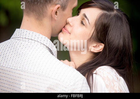 Portrait of young beautiful brunette woman cuddling with her boyfriend, kissing him in cheek with closed eyes and happy smile Stock Photo