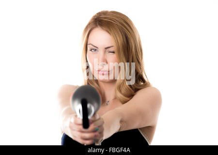 Young funny attractive blond Caucasian woman fooling around, holding black hair dryer like gun pointing towards camera, focus on Stock Photo