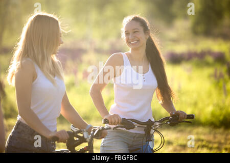 Two young beautiful cheerful women girlfriends wearing jeans shorts on bicycles in park on sunny summer day, having good time, h Stock Photo