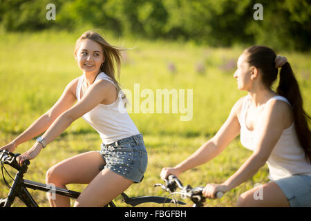 Two cute young happy smiling beautiful women girlfriends wearing jeans shorts riding bikes in park in bright sunlight on summer Stock Photo