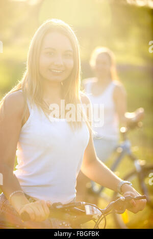 Two cheerful beautiful teenage bicyclist girlfriends wearing casual white tank tops standing with bicycles in park in bright sun Stock Photo