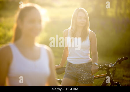 Two attractive girlfriends wearing casual white tank tops and jeans shorts standing with bikes in countryside park on bright sun Stock Photo