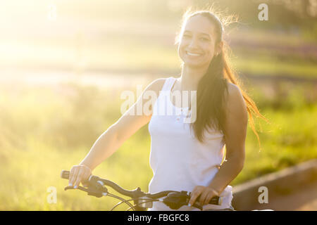 Portrait of happy smiling young asian-caucasian woman posing on bike wearing casual clothes on the road in bright sunlight on su Stock Photo