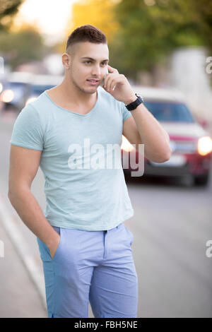Portrait of confident smiling good looking young guy talking on the phone on the street with busy car traffic, young man wearing Stock Photo