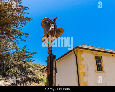A large old rusted metal bell mounted on top of a wooden post stands against a clear blue sky with a deteriorating building. Stock Photo