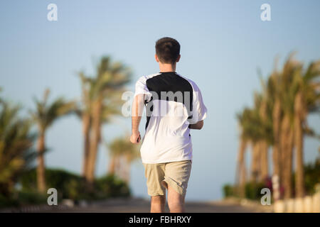 Healthy lifestyle: sporty young man running away from camera, working out outdoors in the summer sunny street in tropics, palm t Stock Photo