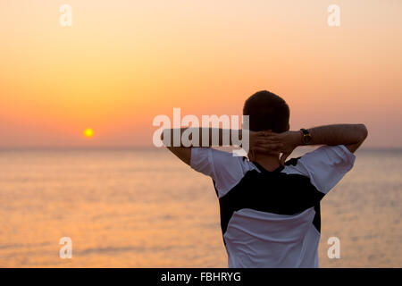 Young man standing at the seashore against colorful sunrise or sunset sky in carefree pose with hands behind his head, enjoying Stock Photo