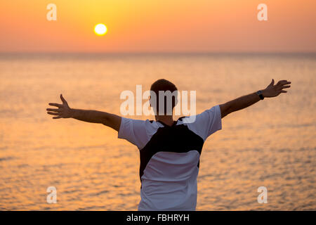 Young man standing at the seashore with raised arms feeling happy, watching colorful sunrise or sunset sky in carefree pose, enj Stock Photo