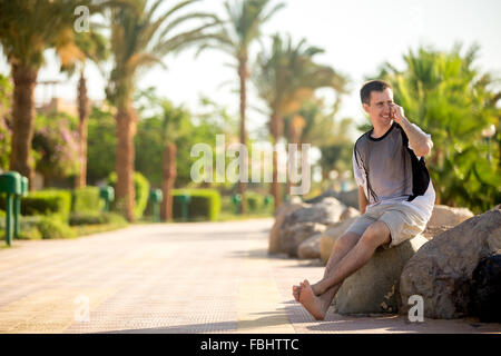 Young handsome happy smiling man sitting barefoot in relaxed pose on the stone on southern sunny street, holding cellphone, maki Stock Photo