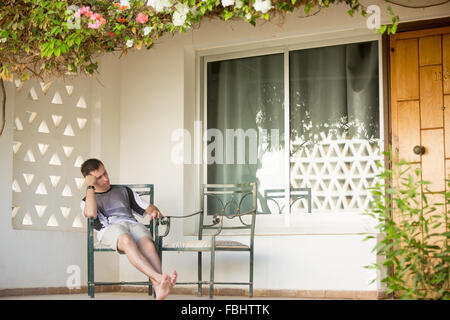 Young handsome man sleeping on chair on the porch of white cottage house covered in flowers Stock Photo