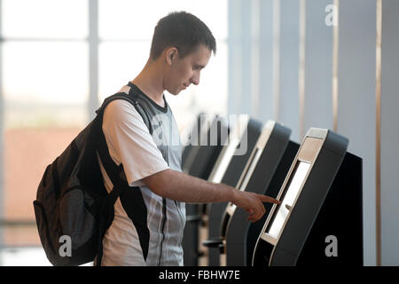 Young man with backpack touching interactive display at self-service transfer machine, doing self-check-in for flight or buying Stock Photo