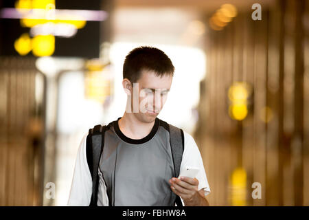 Smiling young man with backpack in his 20s walking in modern building, holding smartphone, looking at screen, using cell phone a Stock Photo