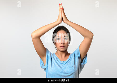 Young woman practicing yoga raising her hands above her head with a serene serious expression as she looks at the camera, upper Stock Photo