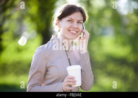 Portrait of happy smiling caucasian woman drinking takeaway coffee, using cellular phone, talking on mobile phone in park Stock Photo