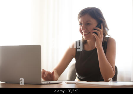 Friendly young office woman in formal wear talking on mobile phone and typing on laptop, taking orders, making business call Stock Photo