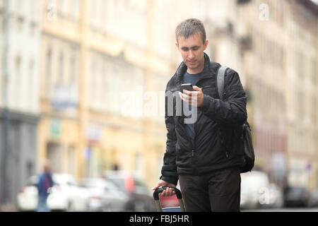 Young smiling attractive man with luggage bag walking in the rainy city street looking at smartphone, using app, gps, searching Stock Photo
