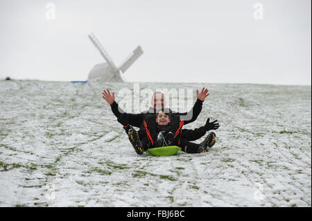 A dad and his son sit on a sledge together and slide down a snow-covered hill In front of one of the Clayton Windmills in Clayton, East Sussex, UK. Stock Photo