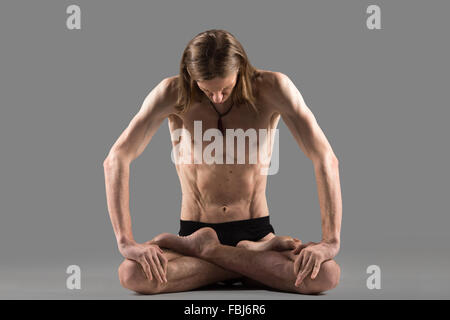 Sporty muscular young yogi man sitting cross-legged in padmasana, lotus posture, studio shot on dark background, full length Stock Photo