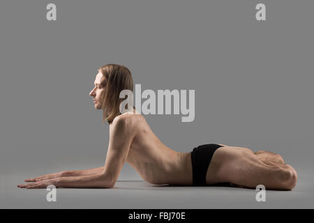 Sporty muscular young yogi man doing backbend exercises, ardha padma bhujangasana, Sphinx Pose with lotus legs, studio shot on d Stock Photo