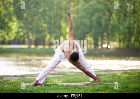 Attractive Indian young yogi man in white linen clothes working out on river bank in park, standing in Cross Triangle posture va Stock Photo