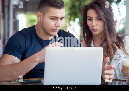Candid portrait of young man and woman in casual clothes sitting in street cafe, using laptop, looking at screen with shocked ex Stock Photo