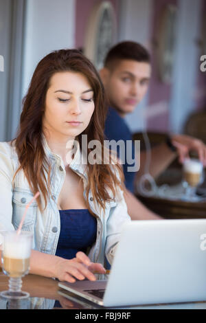 Beautiful serious young woman in casual clothes sitting in street cafe working on laptop while being watched by handsome guy beh Stock Photo