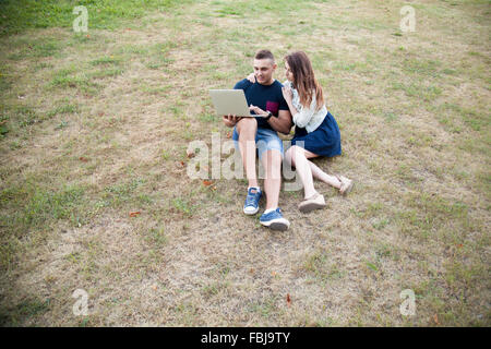 Portrait of smiling handsome young man and beautiful woman in casual clothes sitting on grass in park on summer day, working on Stock Photo