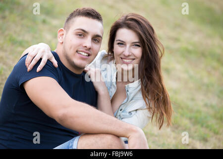 Portrait of beautiful happy smiling young couple on a date embracing, sitting together on grass in park on summer day Stock Photo