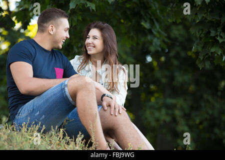 Portrait of beautiful cheerful young couple in love wearing casual clothes relaxing in park, sitting together on the ground on s Stock Photo