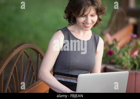 Portrait of happy smiling beautiful young woman sitting on bench on the street in summer, using laptop, looking at the screen Stock Photo