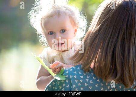 Portrait of happy adorable baby girl with her young mother playing with tree leaf, mom holding her little daughter in arms, walk Stock Photo