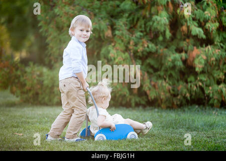 Portrait of two happy cute small siblings playing outside, walking together in park in summer. Smiling brother pushing toy strol Stock Photo