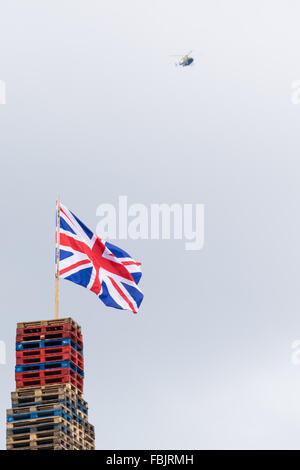 A union jack flag flies from the top of Shankill Road bonfire whilst a PSNI police helicopter soars overhead. Stock Photo