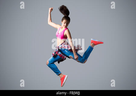 Beautiful young fit modern hip-hop dancer woman working out wearing torn jeans, jumping, studio, gray background, full length Stock Photo