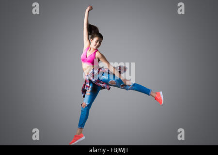 Beautiful young fit modern hip-hop dancer woman working out wearing torn jeans, leap high with joy, studio, gray background Stock Photo