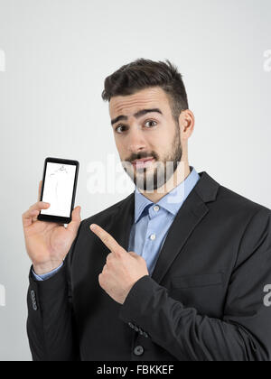Young bearded sad businessman pointing finger at broken smartphone screen. Portrait over gray studio background. Stock Photo