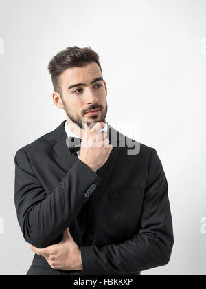 Thinking young bearded man in tuxedo with bow tie touching beard looking away. Desaturated portrait over gray studio background Stock Photo