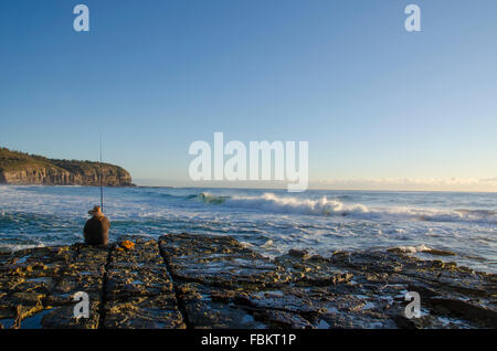 A Caucasian person fishes at dawn at the edge of a rocky platform as waves roll in and the sun rises on a Sydney beach Stock Photo