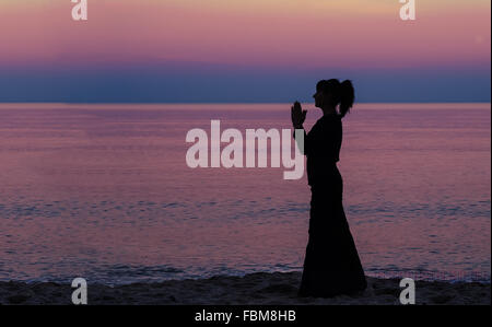 Silhouette of a woman standing on beach with hands in prayer position Stock Photo