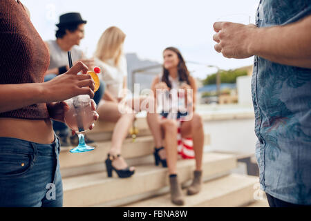 Close up shot of a young people holding cocktails glasses while their friends sitting and talking to each other in the backgroun Stock Photo