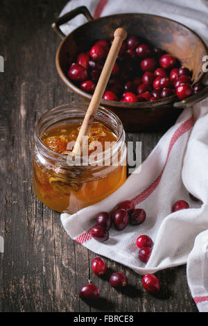 Open glass jar of liquid honey with honeycomb and honey dipper inside and fresh cranberries in vintage bowl over old wooden tabl