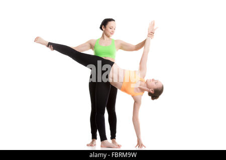 Two beautiful sporty girls practice yoga with partner, coach helps student, doing balance exercise Half Moon Pose, Ardha Chandra Stock Photo