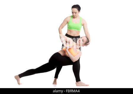 Two beautiful sporty girls practice yoga with partner, coach helps student, stretching, correcting Revolved Side Angle Pose Stock Photo