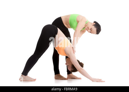 Two beautiful sporty girls practice yoga with partner, instructor assists student, stretching in downward-facing dog yoga pose Stock Photo
