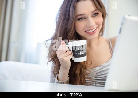 Smiling charming young woman using tablet and drinking coffee from white mug with black area for writing