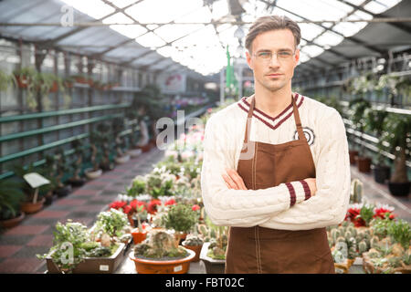 Confident man gardener in brown apron and glasses standing in greenhouse with arms crossed Stock Photo