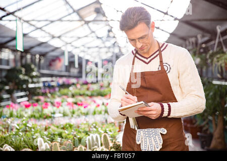 Consentrated male gardener standing and writing in notepad in greenhouse Stock Photo