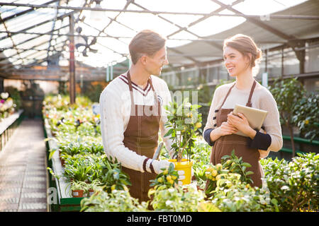 Cheerful woman and man gardeners in brown aprons taking care of plants in greenhouse and using tablet Stock Photo