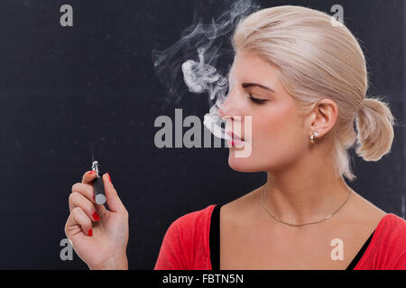 Stylish blond woman smoking an e-cigarette exhaling a cloud of smoke with her eyes closed in enjoyment, profile view on a dark b Stock Photo