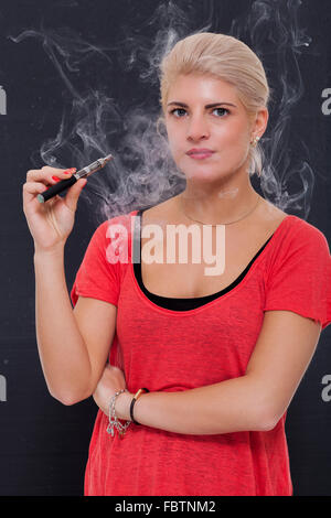 Stylish blond woman smoking an e-cigarette exhaling a cloud of smoke with her eyes closed in enjoyment, profile view on a dark b Stock Photo